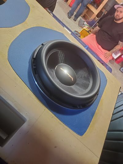 A large black subwoofer mounted in a wooden panel, set against a blue, textured surface. A man is visible in the background.