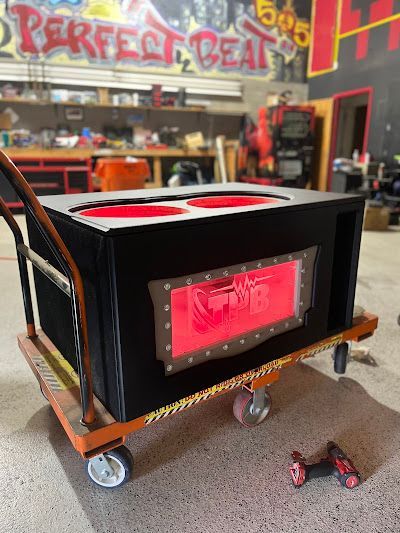A large, black subwoofer box with red accents sits on a hand truck in a garage.