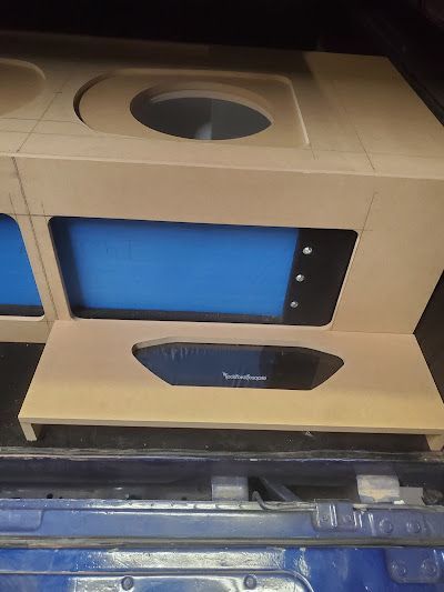 A car audio system's wooden enclosure in a trunk with blue-lit screen, subwoofer openings, and a Rockford Fosgate logo.