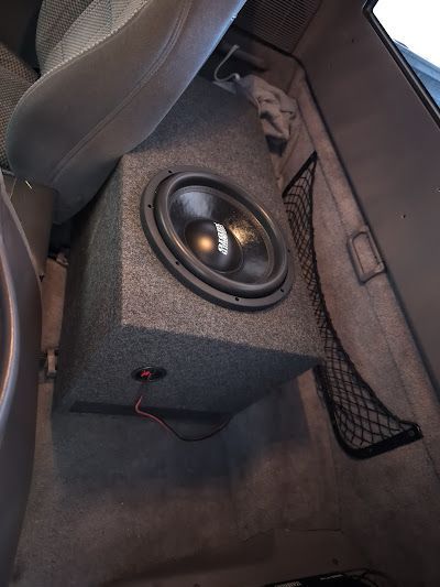 A car subwoofer in a carpeted box sits in a vehicle's footwell. The subwoofer is black and surrounded by gray carpet.