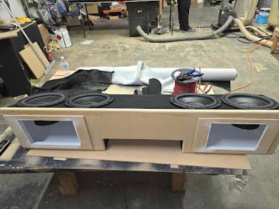 A custom-built speaker box with four subwoofers and two side compartments, placed on a workbench.