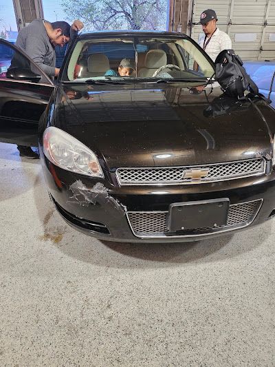 A dark brown Chevrolet Impala with front-end damage being assessed by three people standing around the car in a garage.