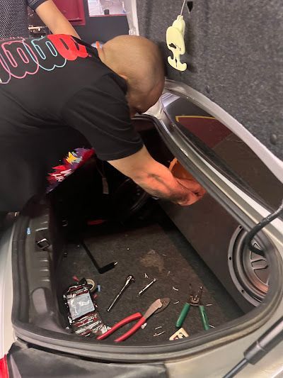 Person working inside the trunk of a car, likely on electrical work. Tools and equipment scattered around.