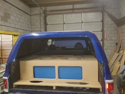 Blue pickup truck bed with a custom subwoofer enclosure installed. Beige wooden box has two blue-faced speaker openings.