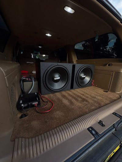 Two large black subwoofers in a car trunk, brown carpet, and wires.