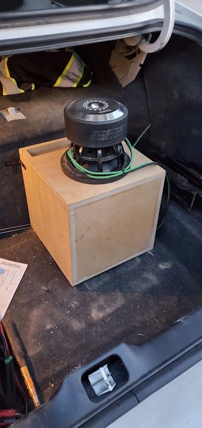 A subwoofer in a wooden enclosure sits inside a car's trunk. The speaker is black with green wiring.