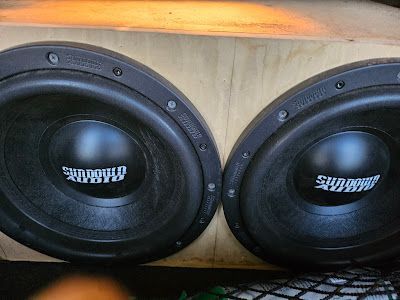 Two black Sundown Audio subwoofers mounted in a light wood enclosure, likely for a car audio system.