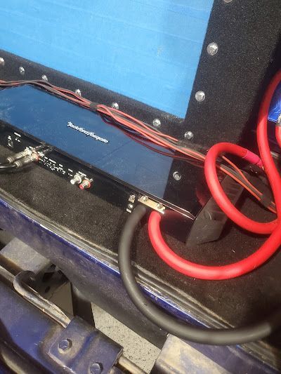 A car audio amplifier with red and black cables connected. It is mounted inside a vehicle.