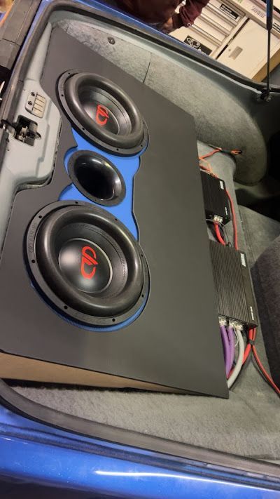 A custom car audio setup in a blue trunk features two subwoofers, a midrange speaker, and two amplifiers.