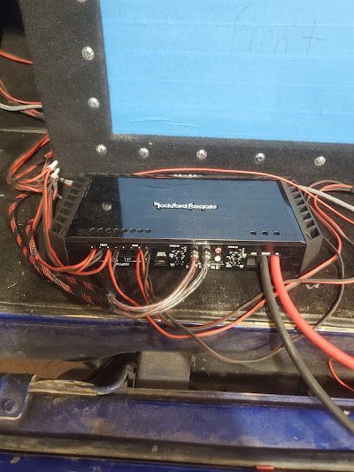 Car audio amplifier with numerous connected wires. Black amplifier on a blue surface, with red and black wires.