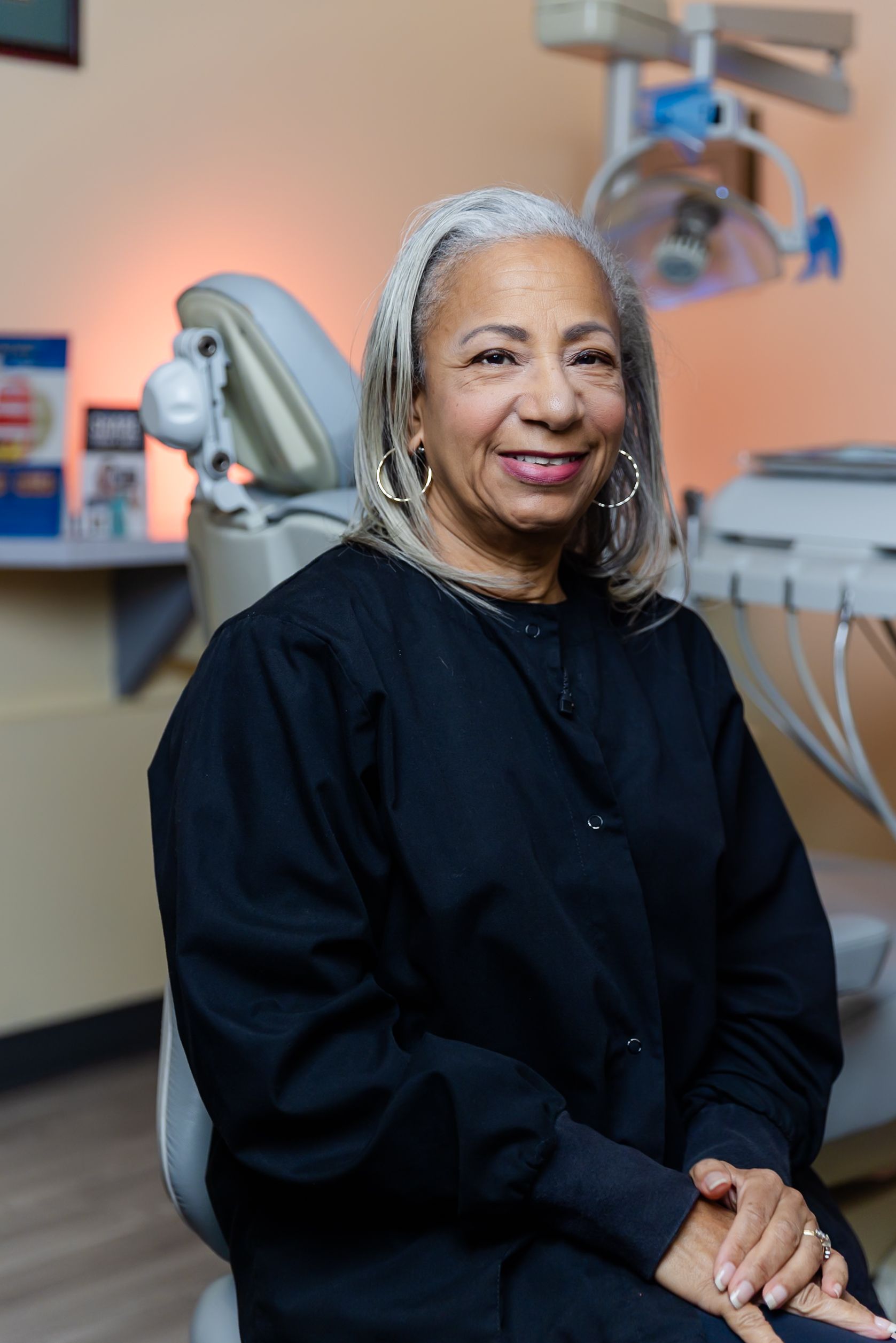 Woman in black scrubs, smiles at camera, seated in a dental chair. Office setting.