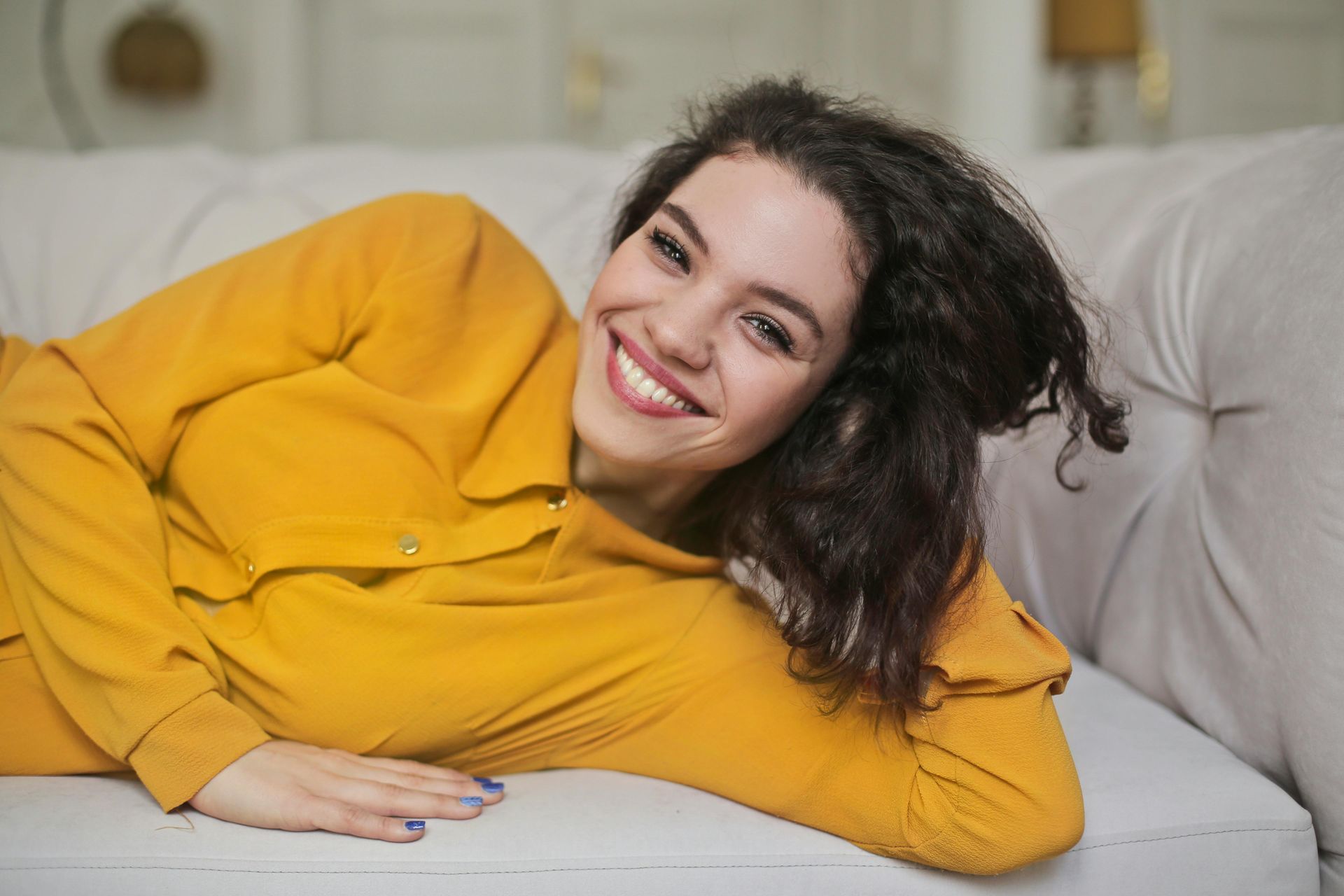 Woman with dark curly hair smiles while lying on a white couch; she wears a yellow shirt.