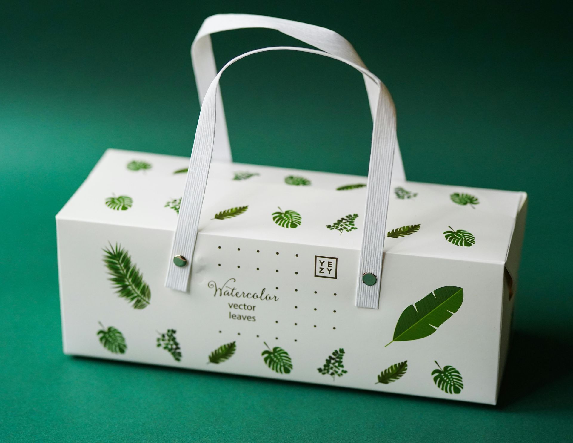 White gift box with green leaf pattern, white handle, against a green backdrop.
