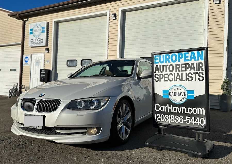 A white bmw is parked in front of a european auto repair specialists sign.