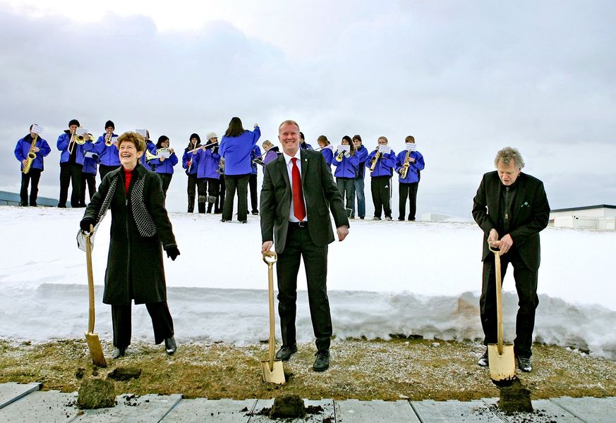 Þrír einstaklingar með skóflur við fyrstu skóflustungu í snjó, með lúðrasveit í bakgrunni.