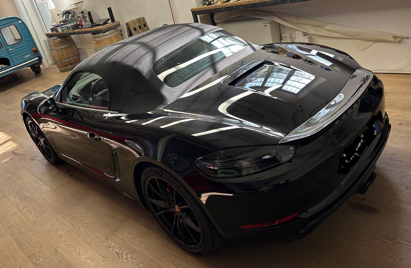 Una Porsche 718 Boxster nera parcheggiata all'interno su un pavimento di legno, vista da una prospettiva posteriore di tre quarti dall'alto.