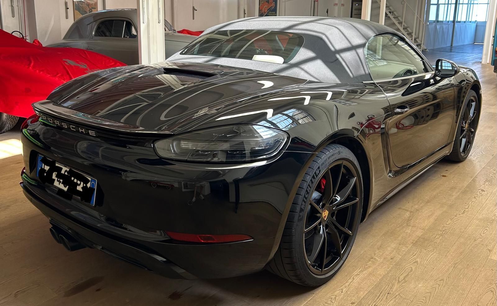Una Porsche 718 Boxster decappottabile nera parcheggiata all'interno di uno showroom, con altri veicoli sullo sfondo.