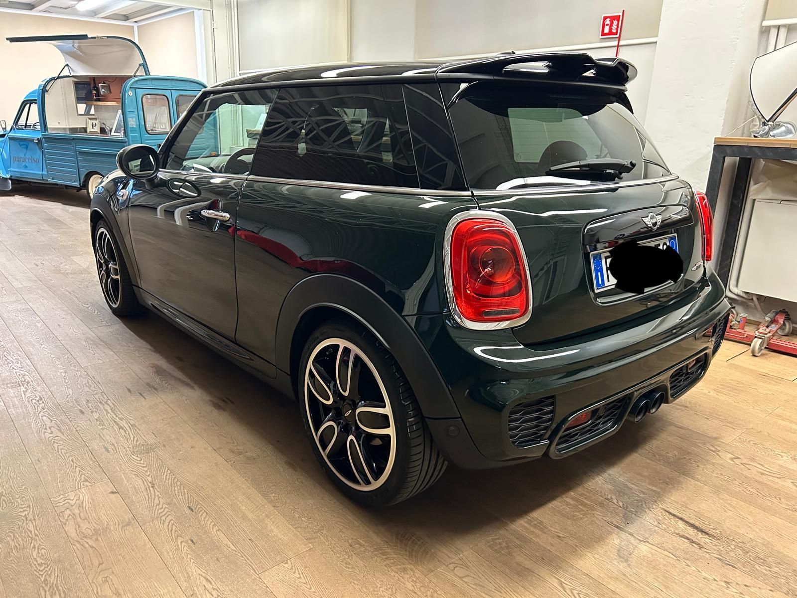Una Mini Cooper a tre porte di colore verde scuro parcheggiata all'interno di un'officina, con un furgoncino per la vendita di cibo di colore azzurro visibile sullo sfondo.