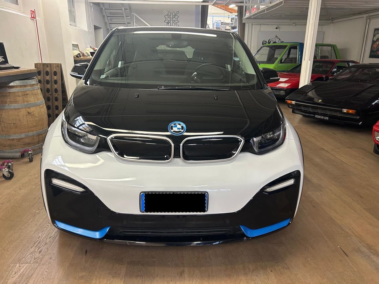 Una BMW i3 bicolore, bianca e nera, parcheggiata in uno showroom insieme ad altri veicoli e a un barile di legno nelle vicinanze.