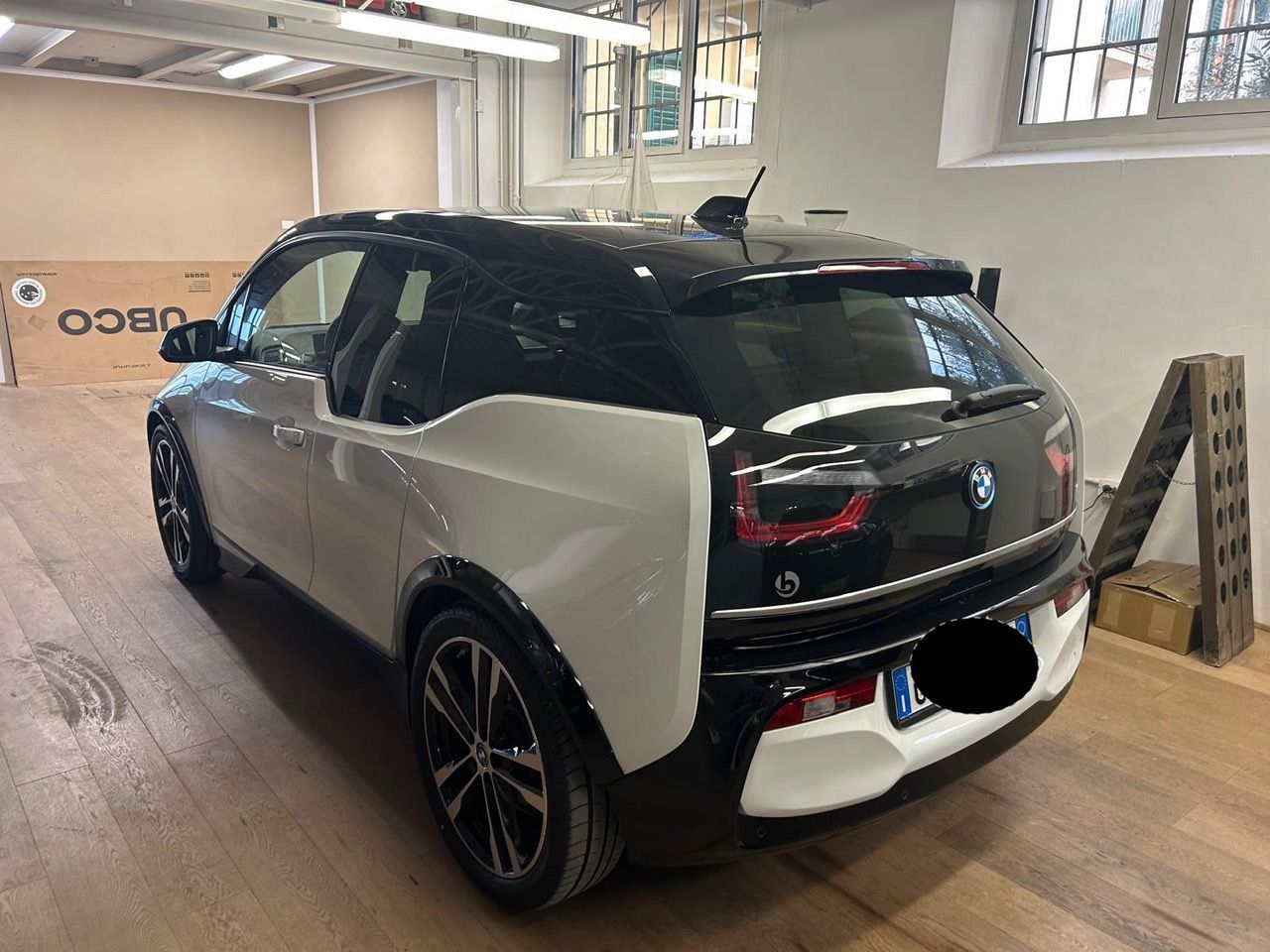 Una BMW i3 bicolore bianca e nera parcheggiata in un interno luminoso, simile a un'officina, con pavimento in legno.