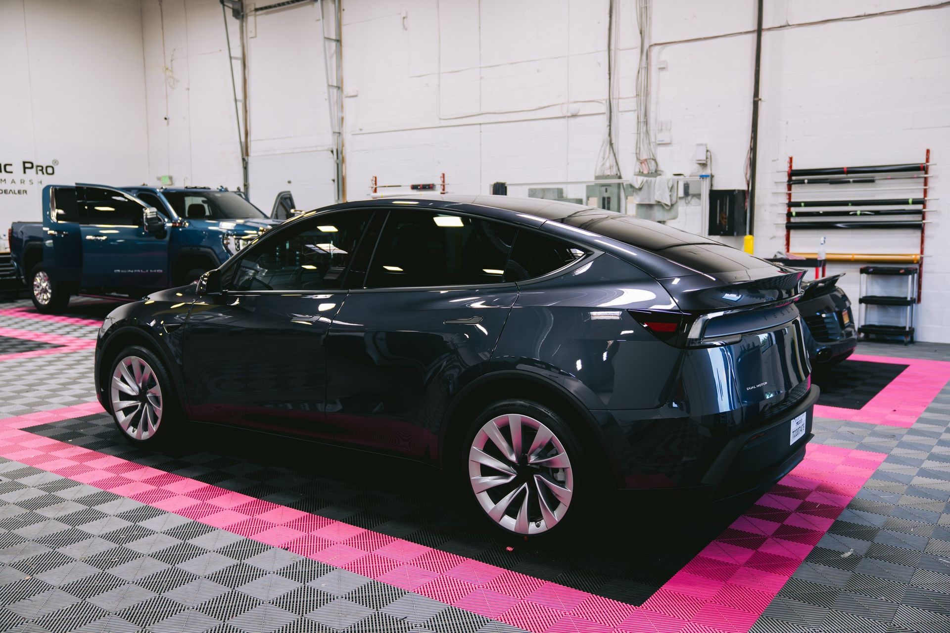 A dark grey Tesla Model Y parked on a patterned workshop floor next to a blue truck.