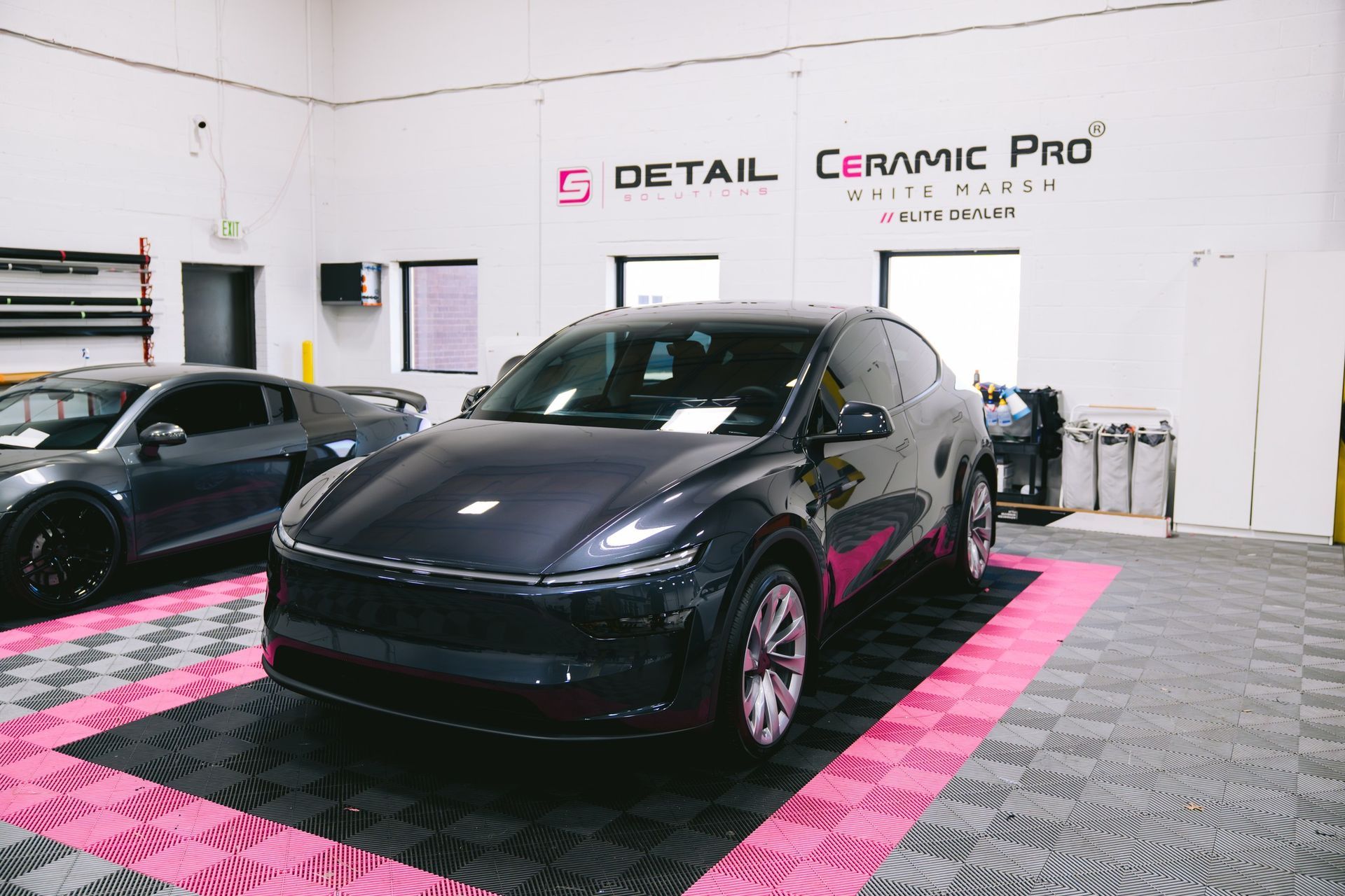 A dark grey Tesla Model Y parked on a checkered floor in a Ceramic Pro automotive detail shop.