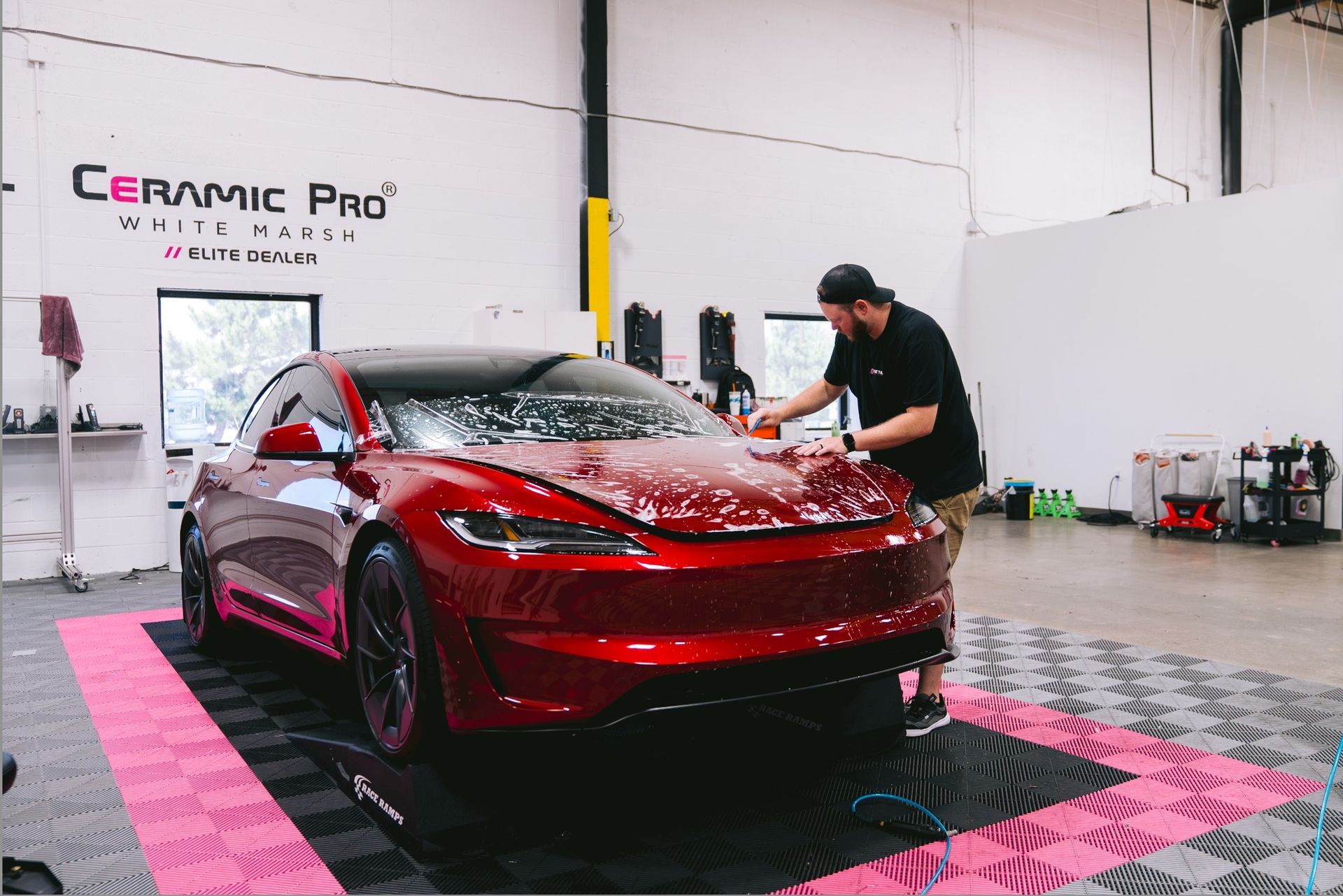 Red car being detailed in a Ceramic Pro shop, with water spray and black/pink floor tiles.