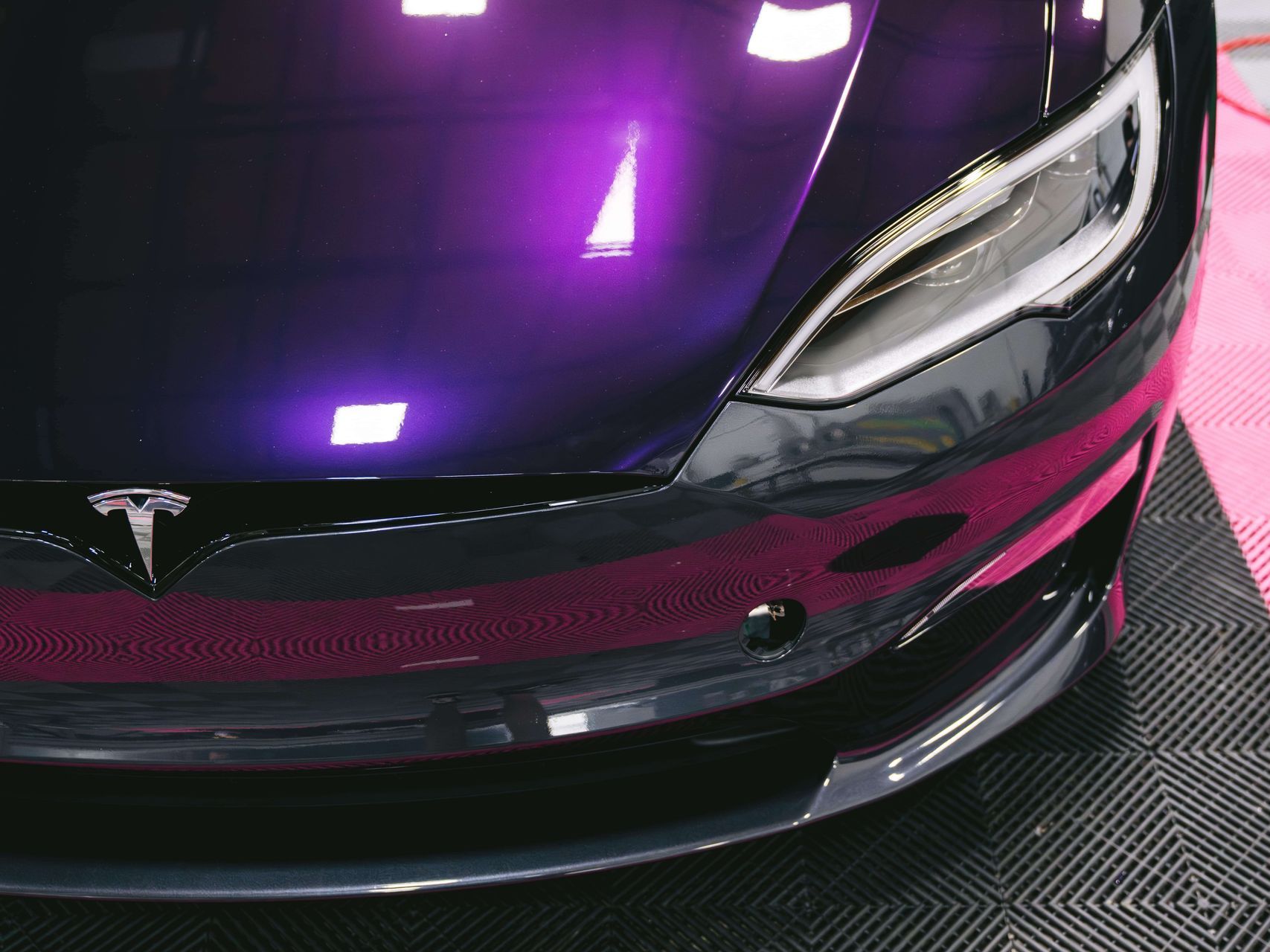 Close-up of a Tesla car with a vibrant purple and black paint job. Shows the front bumper, headlight, and Tesla logo.