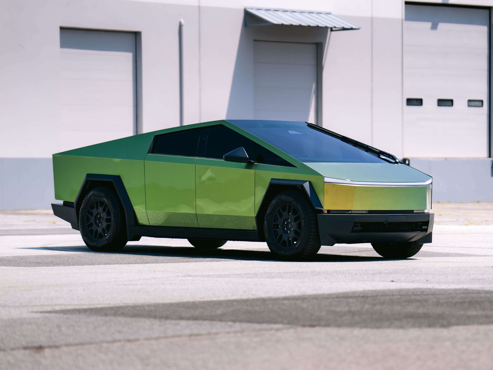 Green Tesla Cybertruck parked in front of a gray building, angled sunlight.