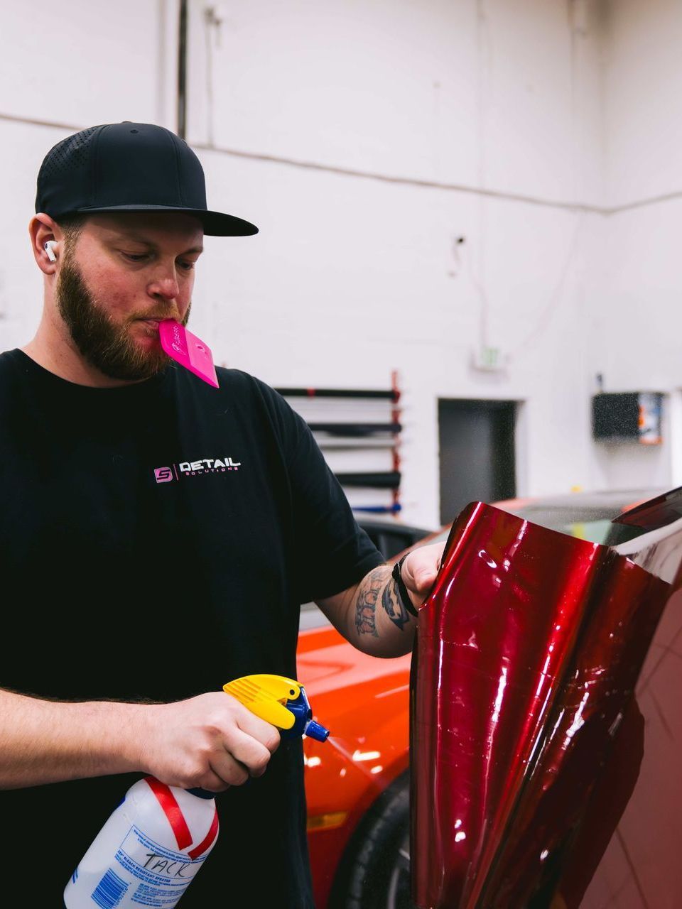 Man spraying car wrap, blowing into a pink tool. Workshop setting with a red car visible.