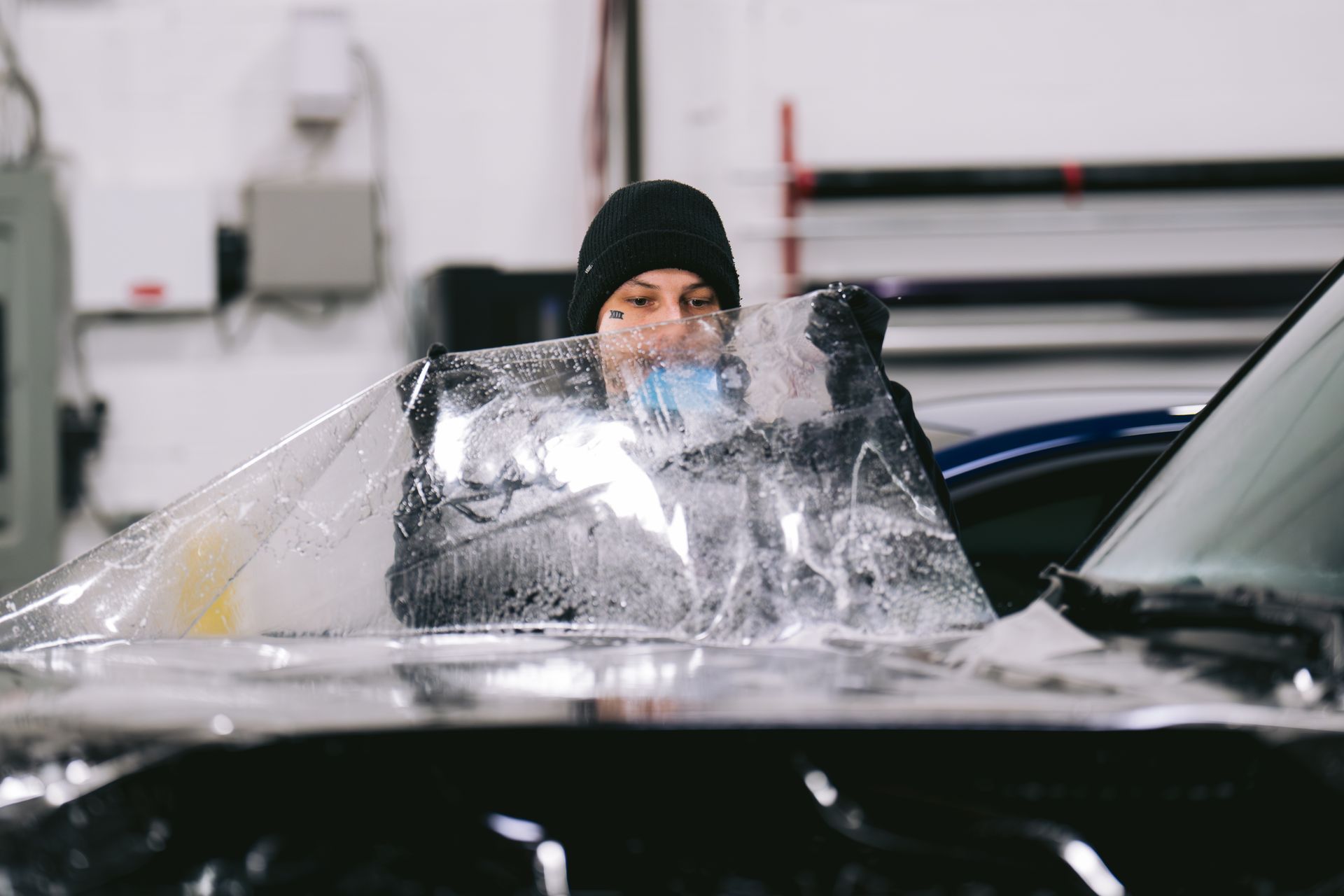 A person wearing a beanie applies a clear protective film to a dark vehicle hood in an auto shop.