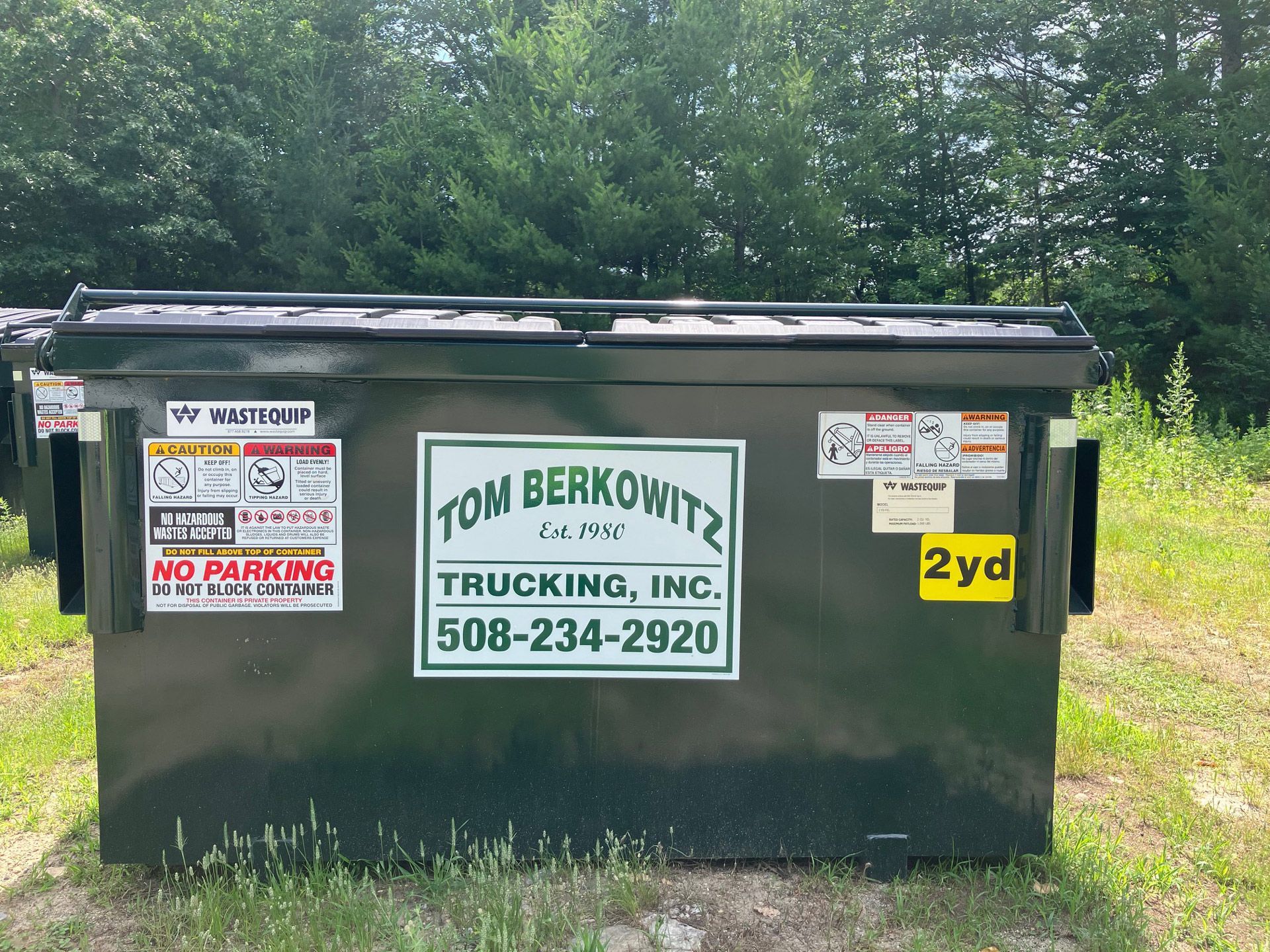 Black dumpster with Tom Berkowitz Trucking, Inc. logo, 2yd capacity, and warning stickers. Outdoors on grassy area.