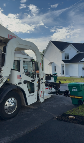 A white garbage truck with a robotic arm lifts a green bin in front of a house on a sunny day.