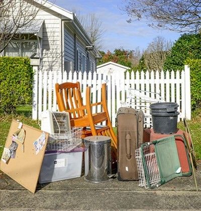 Pile of discarded household items, including a chair, trash cans, and luggage, set out for garbage pickup on a curb.