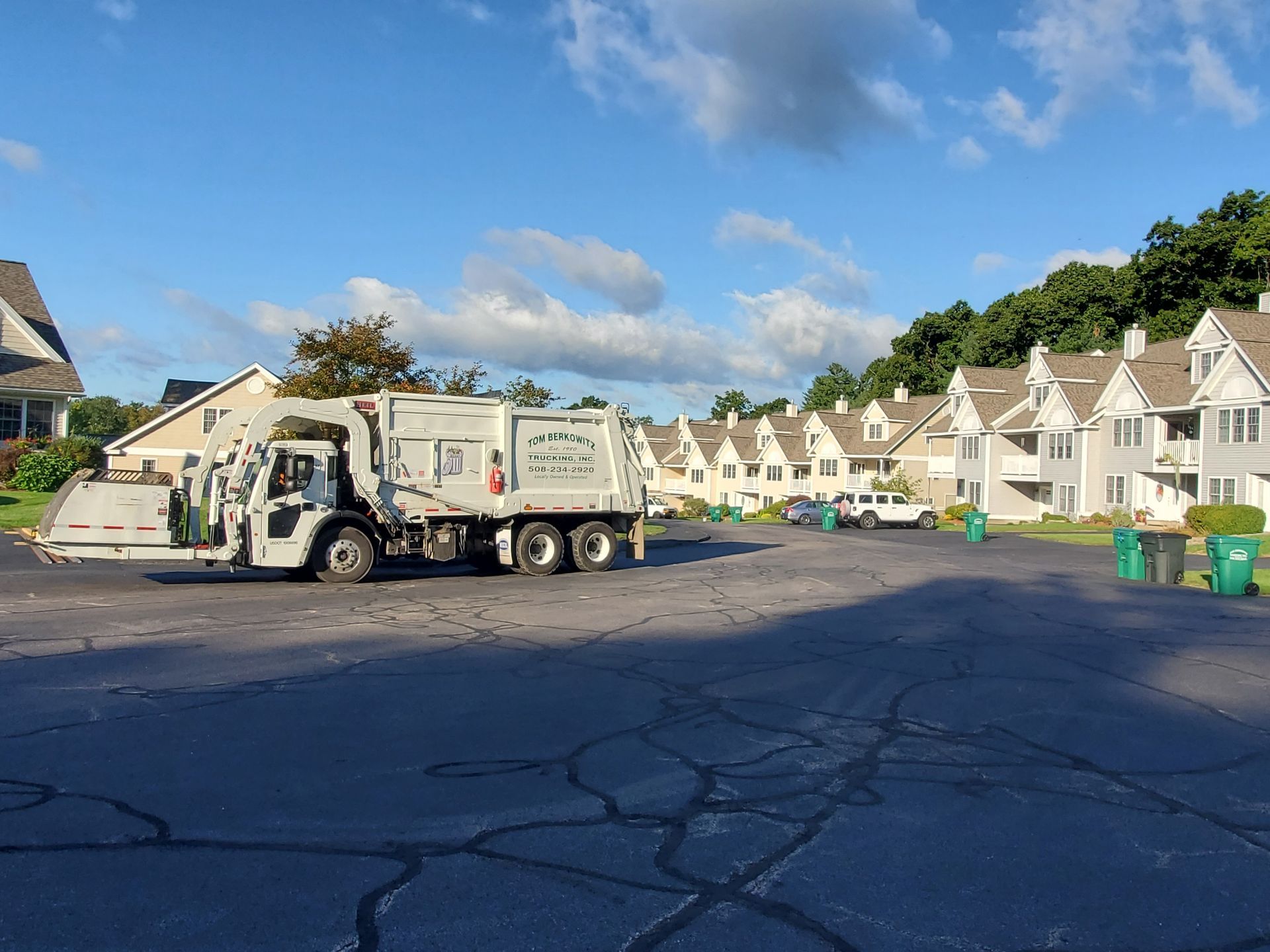 Garbage truck in a residential area, parked next to recycling bins; apartment buildings in background.