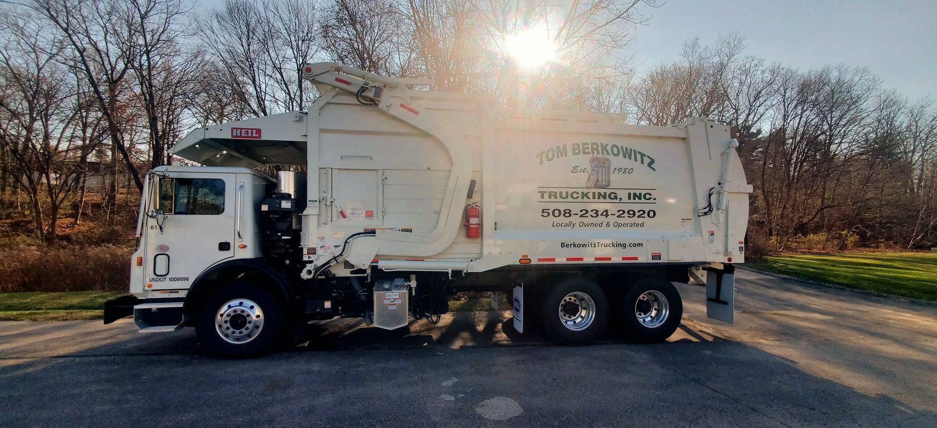 White garbage truck parked outdoors on asphalt, sun shining behind it.