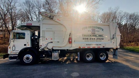 White garbage truck on a paved road, parked. Trees in the background, sun shining.