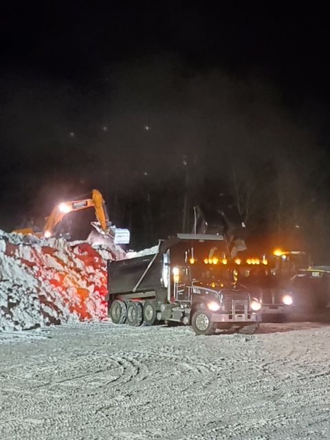 Excavator loading a dump truck at night. Snow-covered area, trees in the background, lit by vehicle lights.