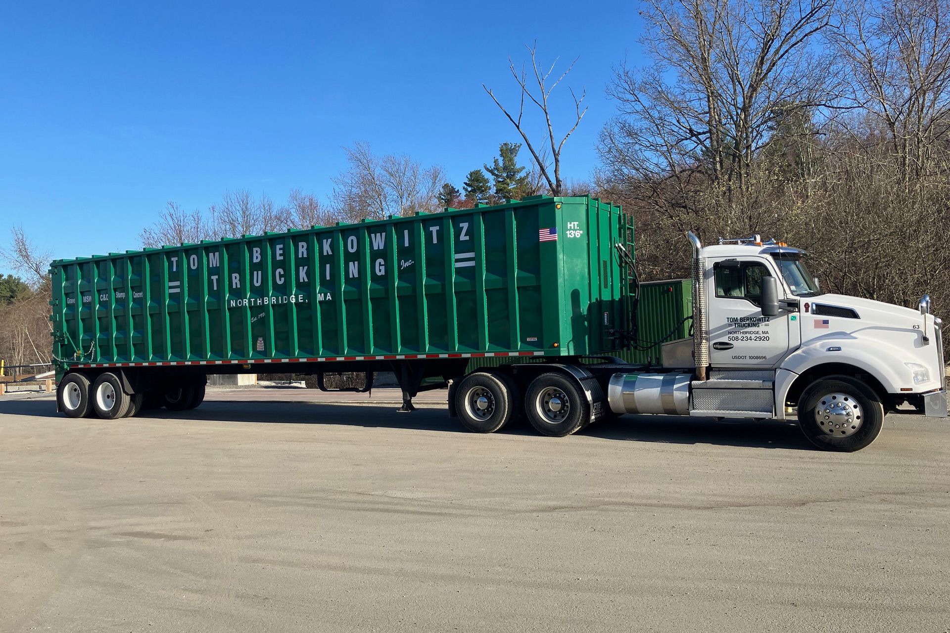 White semi-truck hauling a long, green container on a paved surface on a sunny day.