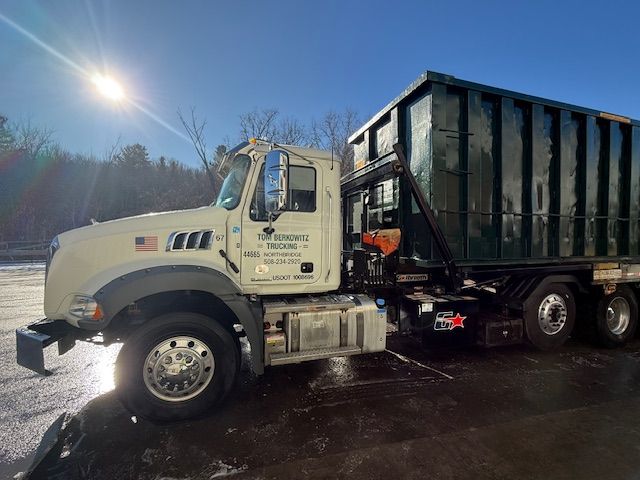 Row of green dumpsters on a gravel surface next to trees.