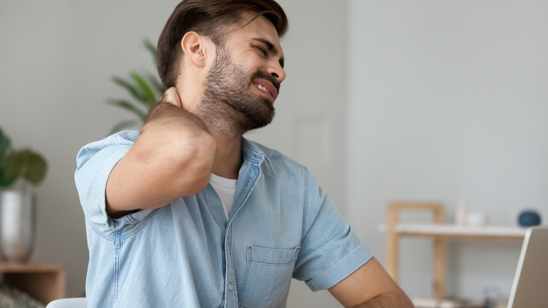 Man with beard clutches his neck, showing discomfort; indoor setting.