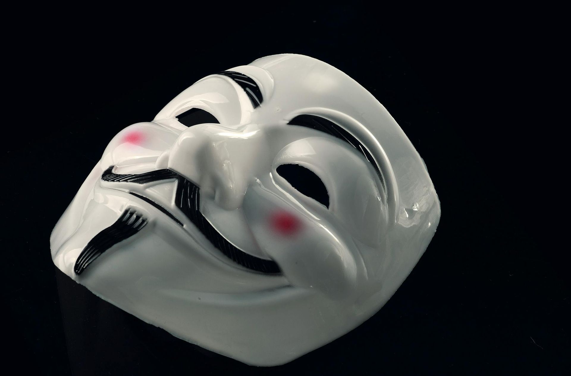 White Guy Fawkes mask with a smile, black eyebrows, and pink cheeks against a black background.