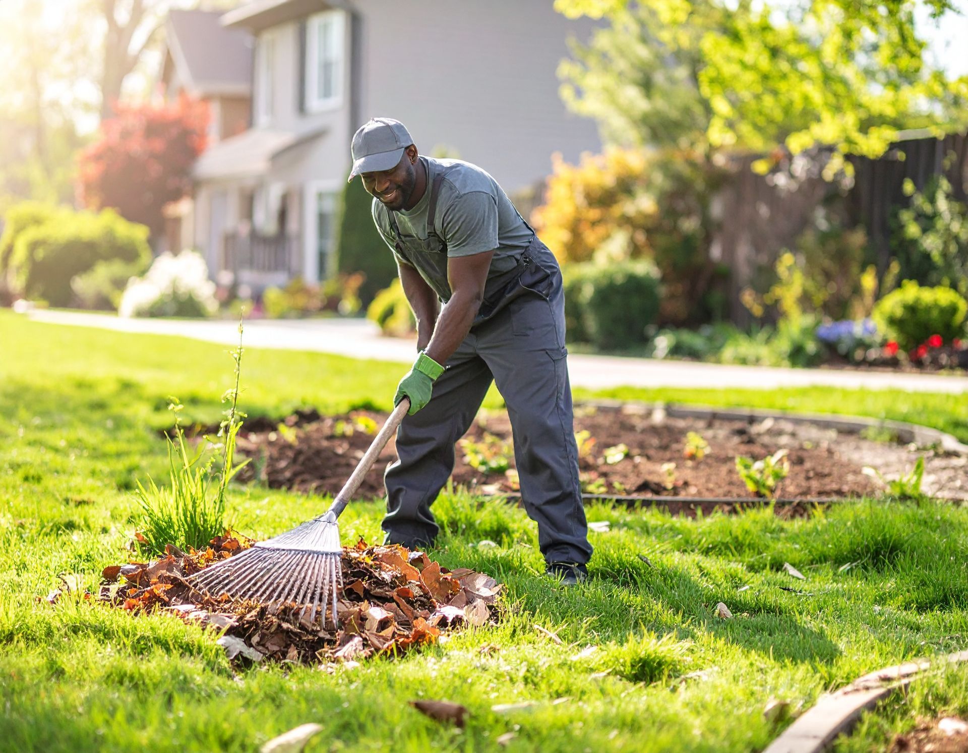 A person wearing work clothes and a cap rakes dirt in a grassy yard, smiling while working.