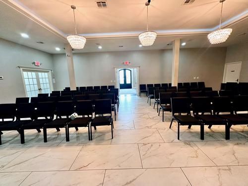 Rows of black chairs face a center aisle in a bright room with chandeliers, doors, and a marble floor.