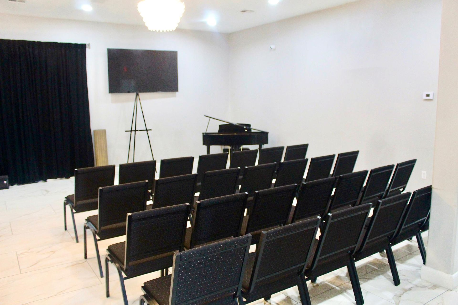 Rows of black chairs face a grand piano, screen, and stage with black curtain, in a white room.