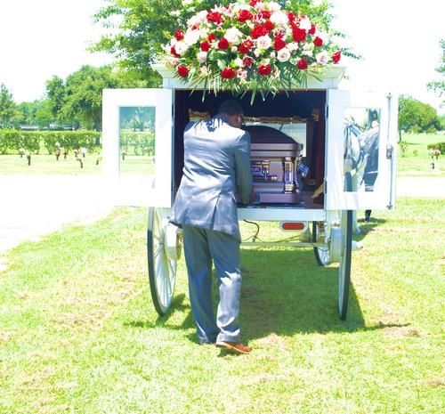Man in suit placing casket from flower-laden white carriage at cemetery.