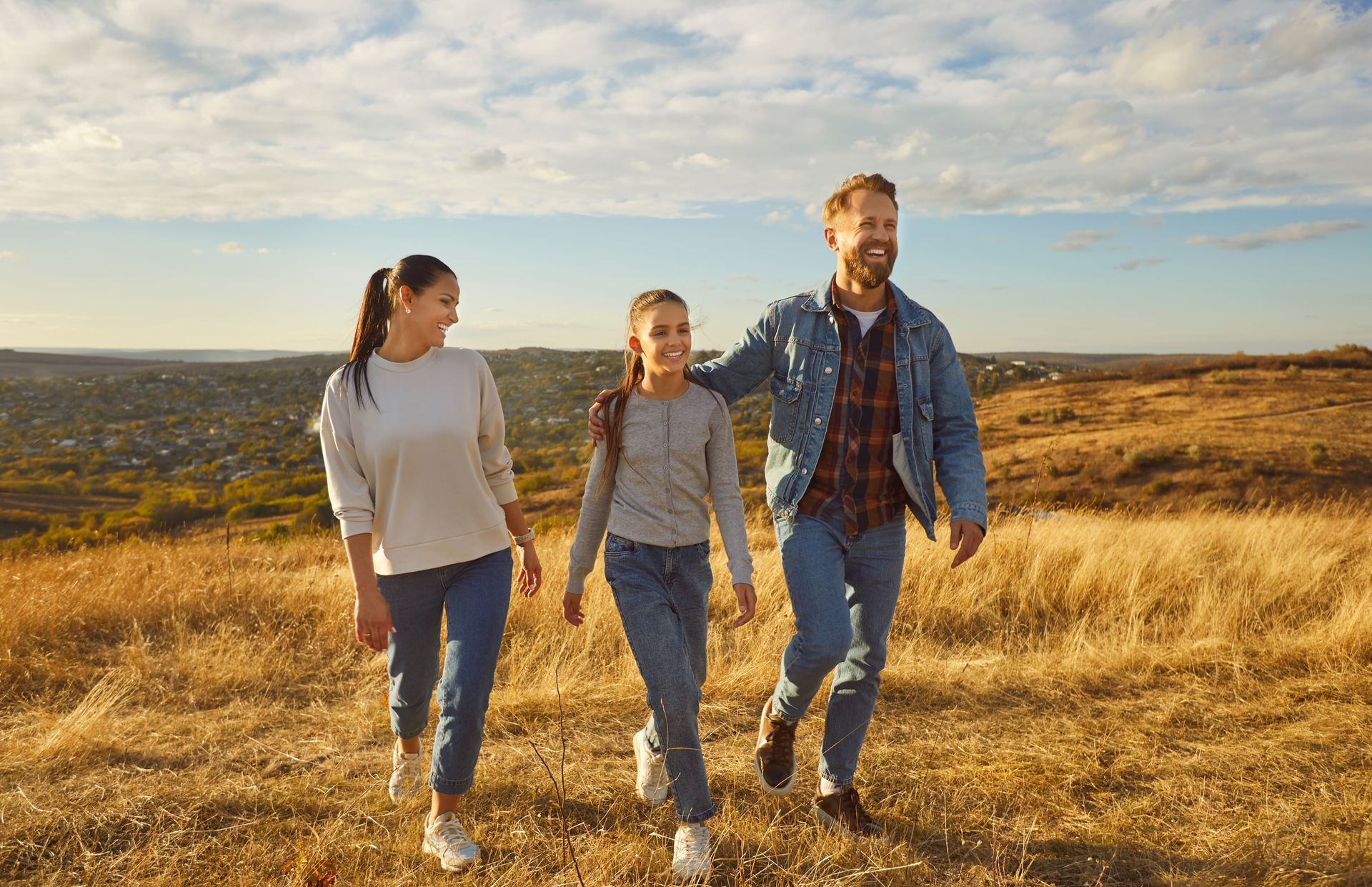 Family walking in a field: mother, daughter, father with arm around daughter, smiling, sunny day.