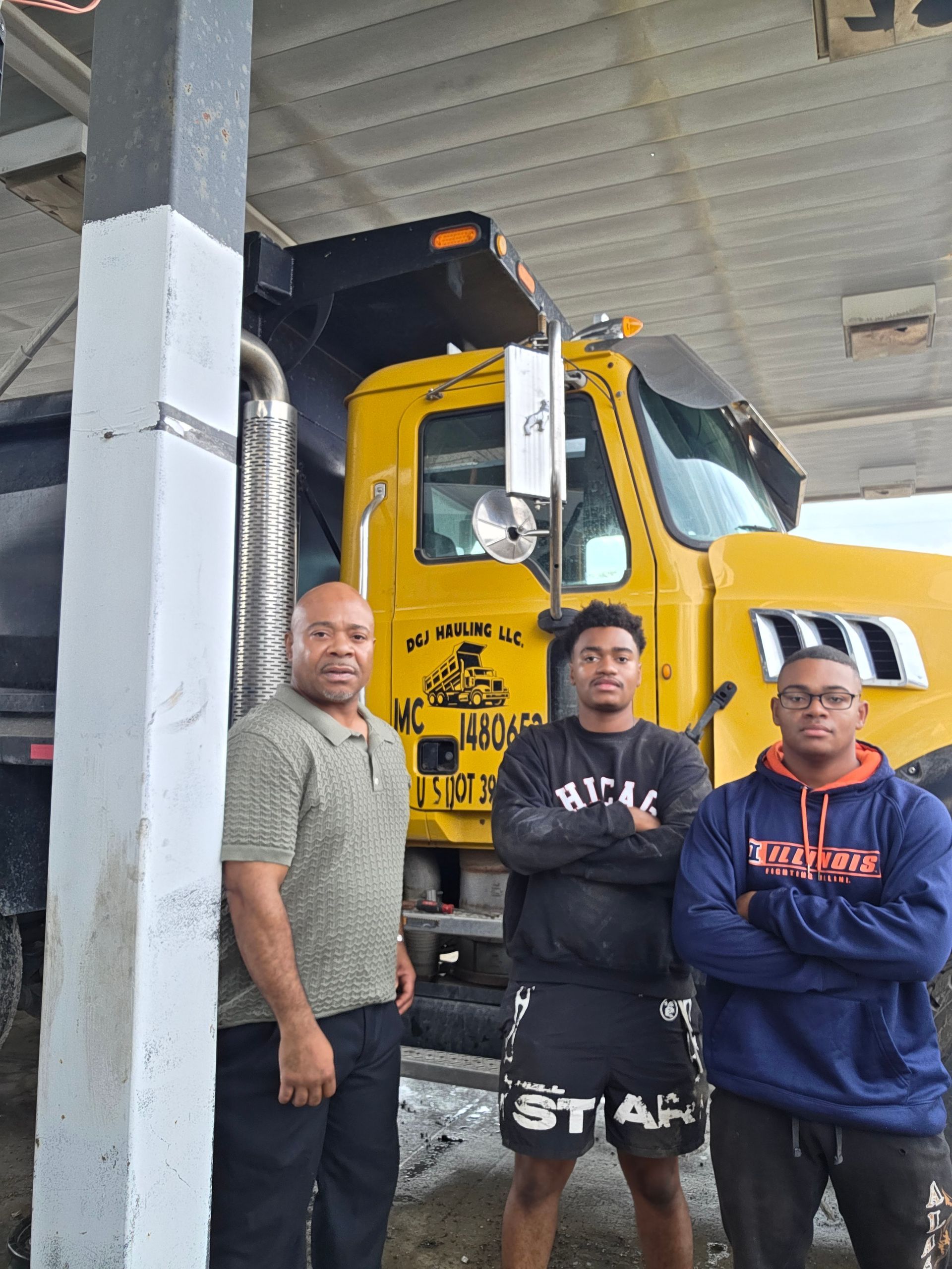 Three men stand in front of a yellow dump truck. One leans on a column; two have arms crossed.