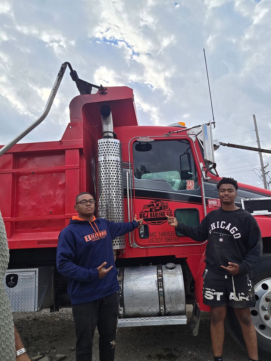 Two people pointing at a red dump truck in an outdoor setting with a cloudy sky.