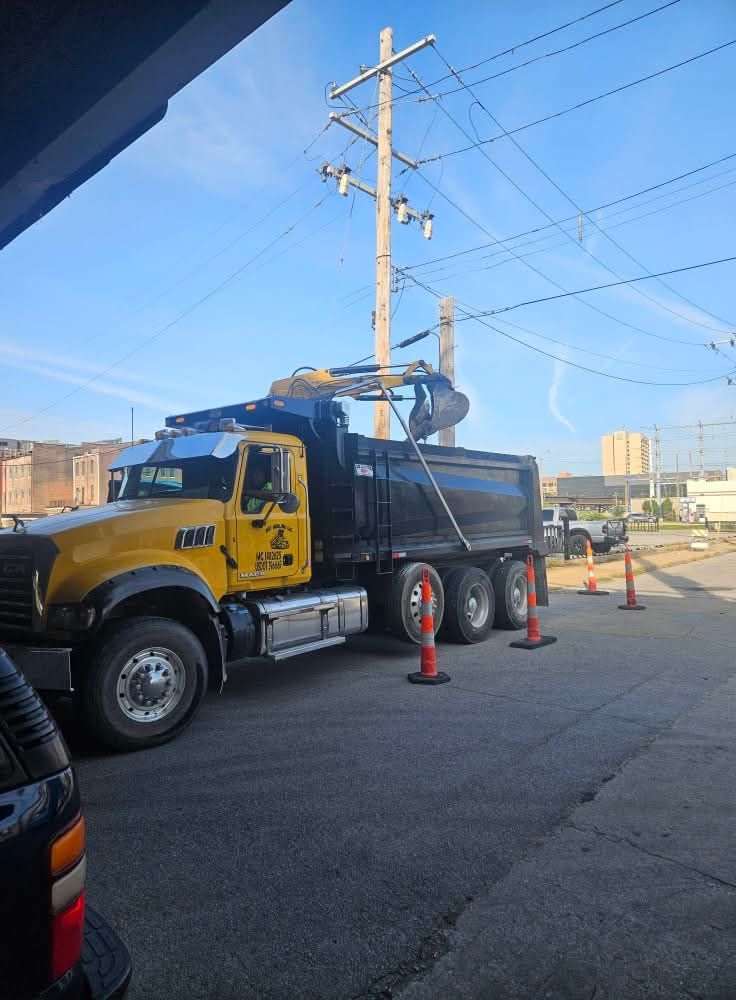 Yellow dump truck with a black bed loading debris near power lines and construction cones.